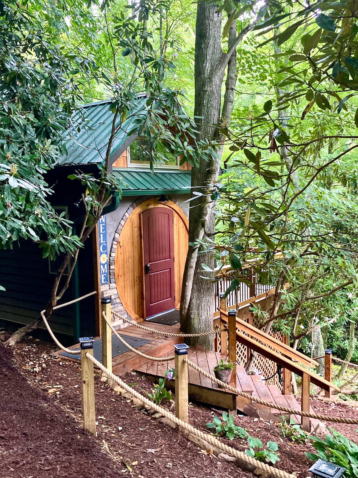 A unique cabin entrance features a circular wooden door framed by lush greenery. A sloping pathway is lined with a rope railing, leading to a set of wooden stairs that descend to the cabin, surrounded by trees and vibrant foliage.