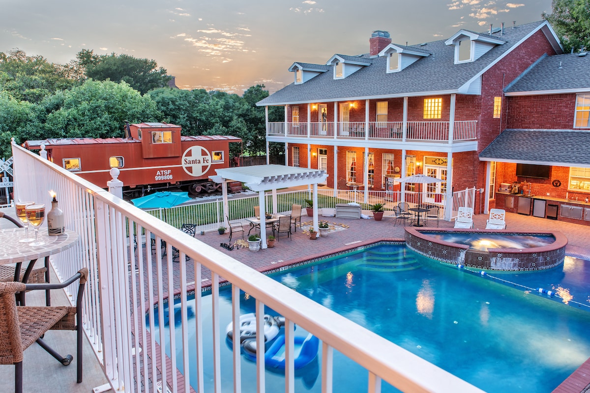 The image displays a vibrant outdoor space featuring a pool and hot tub surrounded by lush greenery. A converted Santa Fe caboose is positioned nearby, with a two-story brick building in the background. A patio with a shaded seating area offers a view of the scenery.