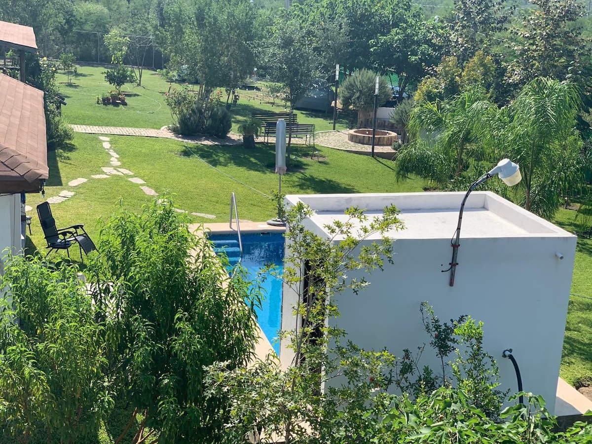 A clear view of a well-maintained backyard features a sparkling pool surrounded by green grass and scattered trees. A pathway leads through the garden, while a cozy seating area and a fire pit are visible in the distance, inviting outdoor relaxation.