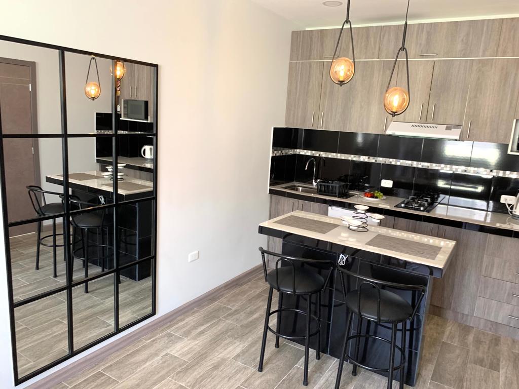 A modern kitchen area is featured, showcasing sleek black cabinetry and stainless steel appliances. A dining table is set for two, accompanied by high-backed chairs. A large, decorative mirror reflects the space, enhancing its brightness and openness.