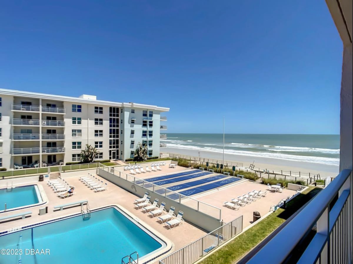 An ocean view is presented from the balcony, showcasing two Olympic-sized swimming pools surrounded by lounge chairs. A well-maintained condominium complex is visible in the background, with its modern façade standing against a clear blue sky and ocean waves gently crashing in the distance.