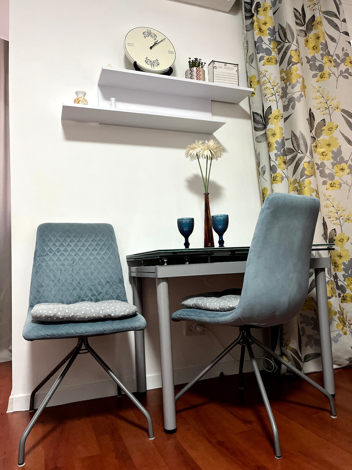 A small dining area is presented with a sleek table and two upholstered chairs. Decorative items are arranged on a nearby shelf, including a clock and a vase. A curtain with floral patterns enhances the space, while warm wooden flooring provides a cohesive backdrop.