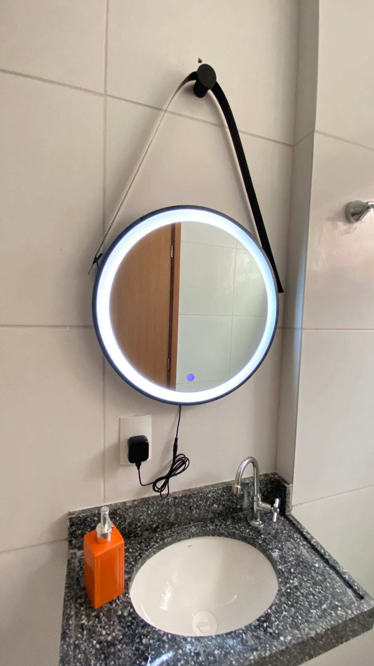 A circular mirror with a built-in light ring is hung on a white tiled wall, above a polished granite countertop. An orange soap dispenser sits beside a modern faucet, contributing to a clean and sleek bathroom design.
