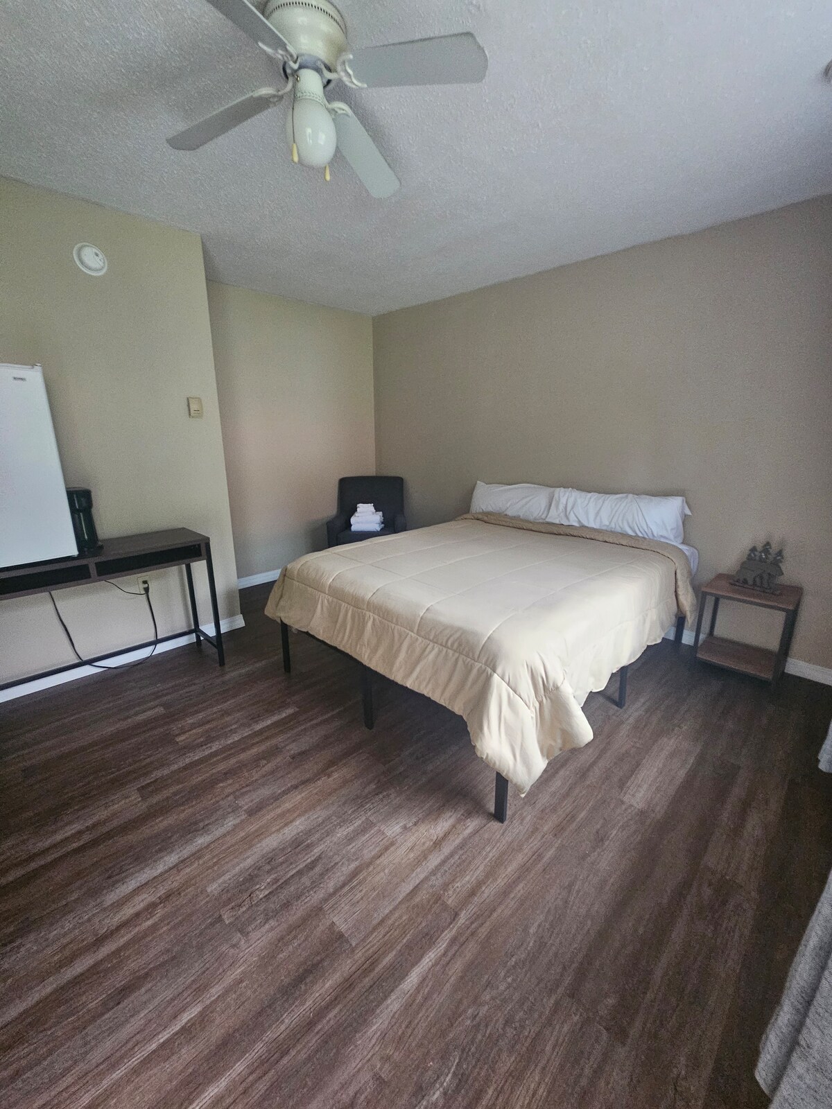A spacious room features a comfortable bed with a light-colored duvet and crisp white linens. A small desk and chair are positioned against one wall, alongside a compact refrigerator. Soft natural light filters through the room, accentuating the wooden flooring.