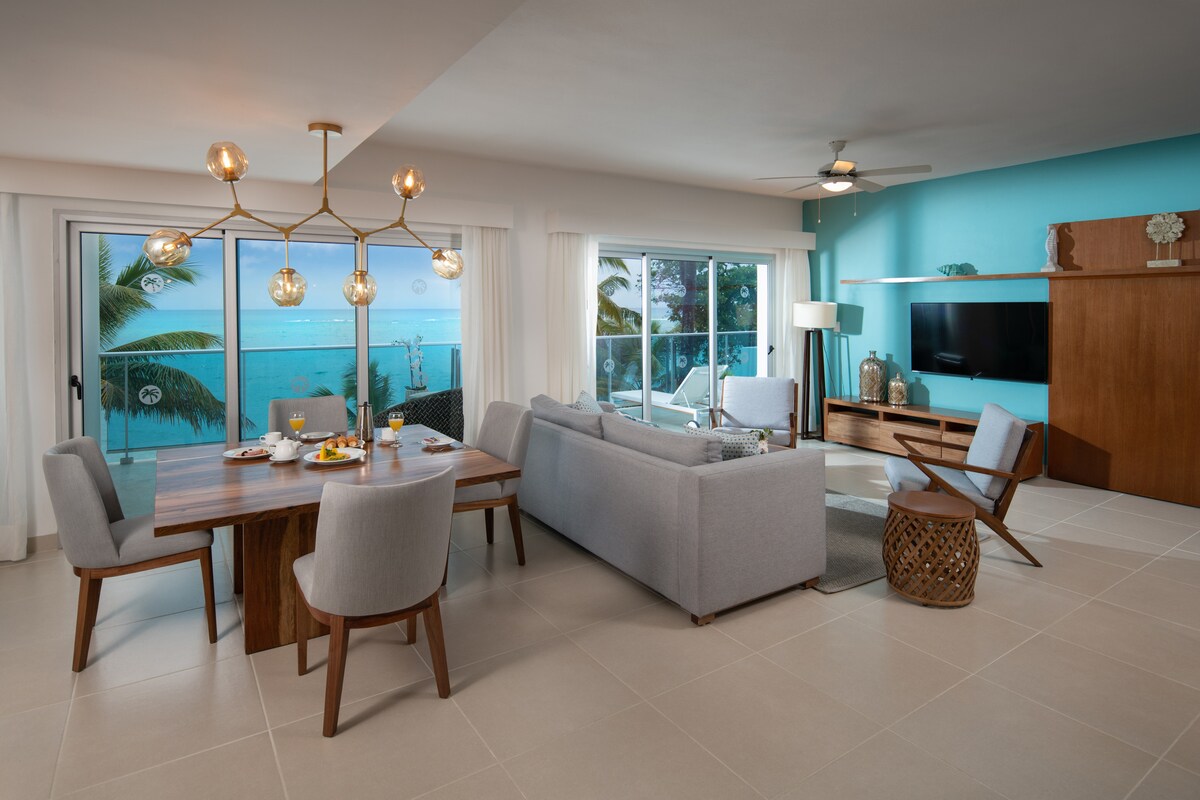 A bright living and dining area showcases a modern design with a blue accent wall. A wooden dining table is set for four, while a comfortable sofa faces a mounted television. Large glass doors provide views of the turquoise ocean and allow natural light to fill the space.