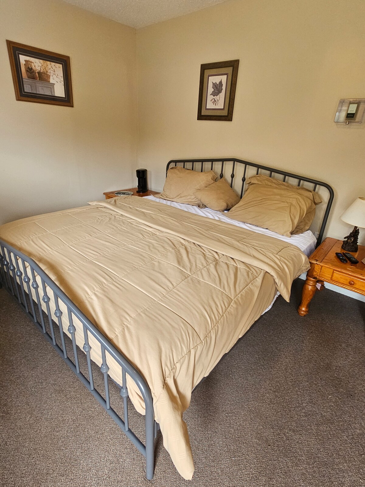 A compact sleeping area features a metal bed frame with a neatly made bed layered in a light beige comforter. Two pillows rest against the headboard. A small nightstand is positioned beside the bed, holding a lamp and a decorative item.