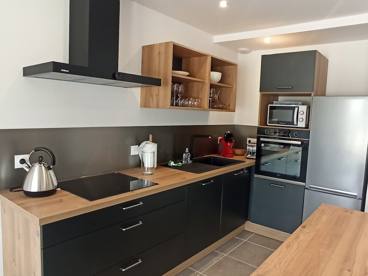 A modern kitchen is presented with sleek black cabinetry and light wood shelving. Equipped with a kettle, coffee maker, and various appliances, the space features a flat cooktop and an oven. The neutral tiles offer a clean look, enhancing the kitchen's functionality.
