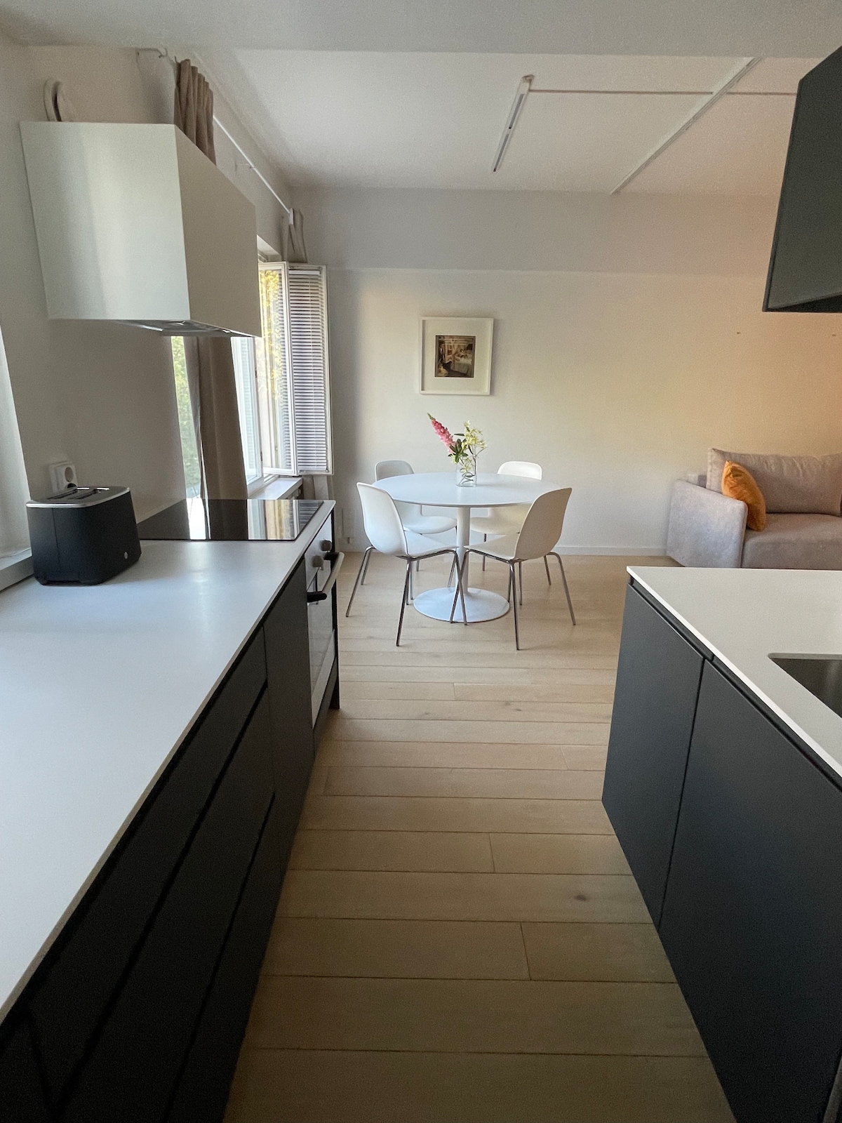 An open kitchen area features modern black cabinetry and a light countertop. A small round dining table with four white chairs is positioned in the corner, adorned with a vase of flowers. Natural light enters through the window, illuminating the adjacent light-colored living space.