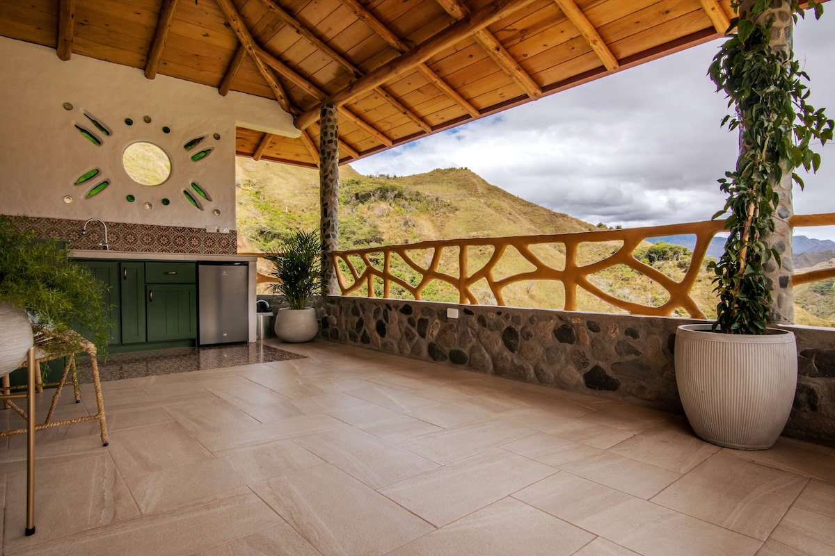An outdoor kitchen area features a rustic design, with a stone countertop and green cabinetry. Large windows offer sweeping views of the surrounding Andean mountains, and decorative details include a circular window and potted greenery. The wooden ceiling adds warmth to the space.