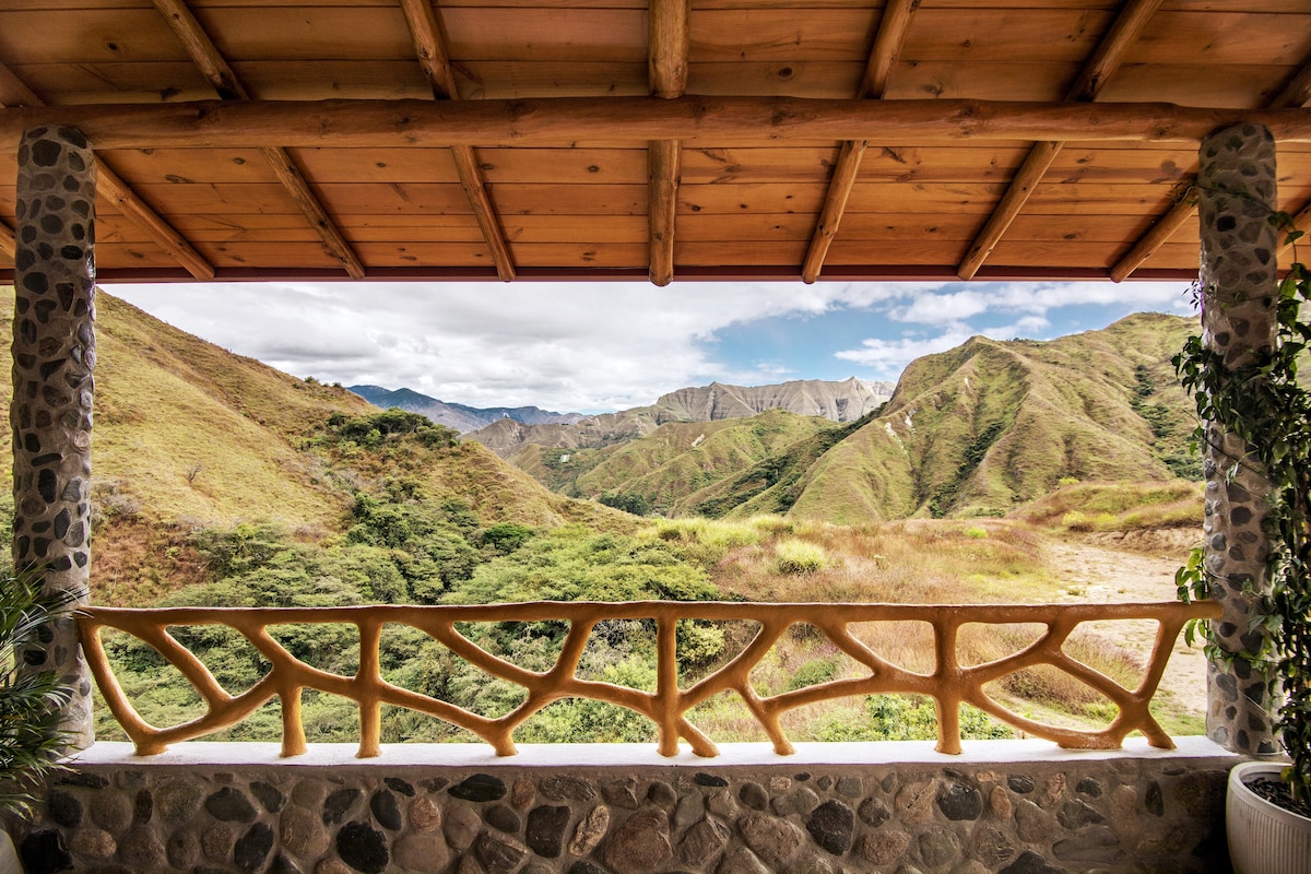 A scenic view of rolling green hills and mountains is framed by a natural stone railing. The wooden overhang provides shade, enhancing the landscape's natural beauty while inviting guests to enjoy the tranquility of the surrounding nature.