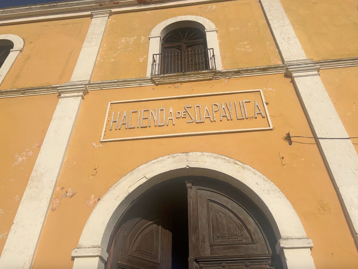 The exterior of the hacienda is highlighted by a warm ochre facade, showcasing elegant white architectural details. A prominent wooden entrance door features intricate carvings, inviting guests in. Above the door, the name 'Hacienda de Soapayuca' is displayed in a classic font, framed by a decorative arch.