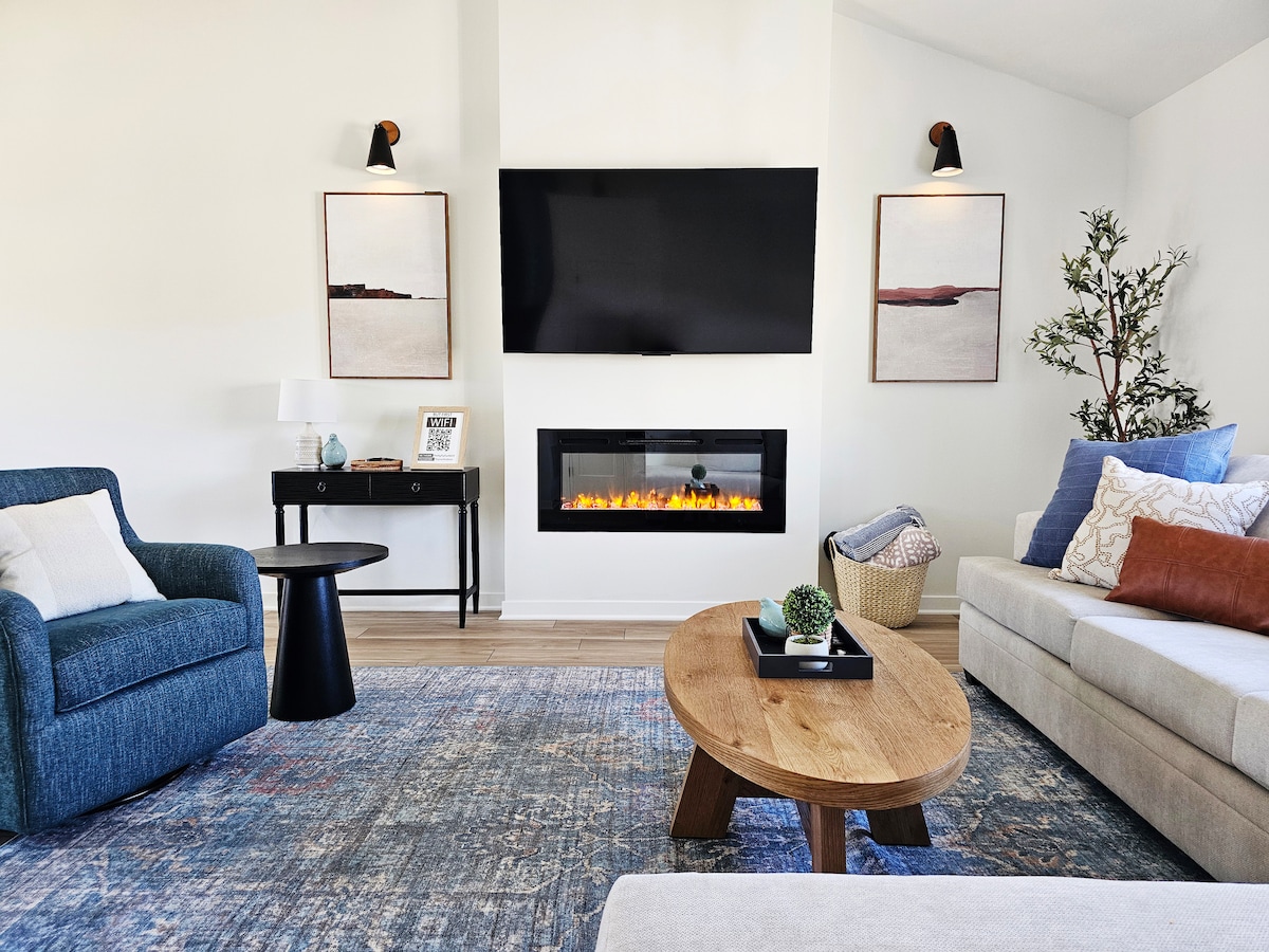 A welcoming living area features a comfortable sectional sofa and an accent chair, accompanied by a round wooden coffee table. An electric fireplace is set into the wall below a mounted TV, with framed artwork hanging above, enhancing the bright, neutral atmosphere.