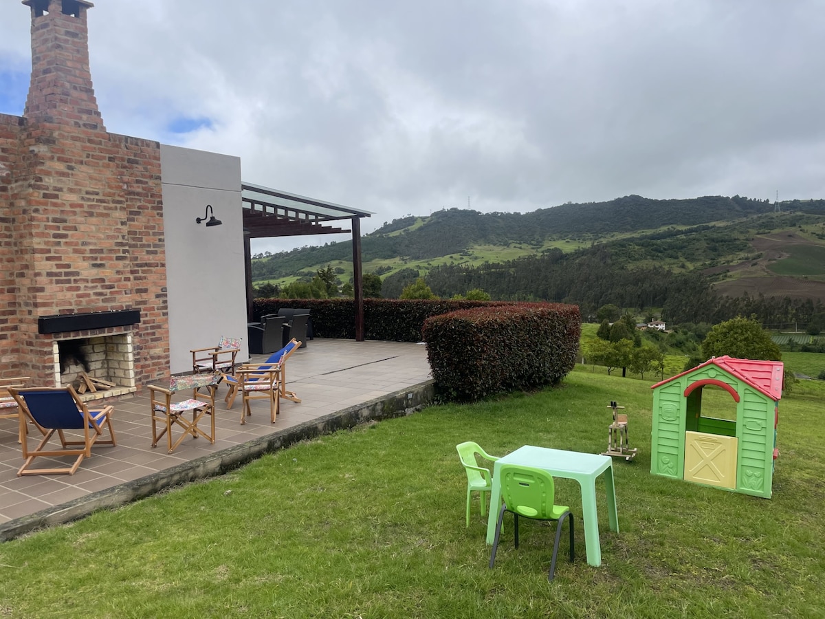 An outdoor area is presented with comfortable seating and a built-in barbecue. A small children's playhouse and a brightly colored table and chairs set are positioned on the manicured lawn, with scenic hills visible in the background.