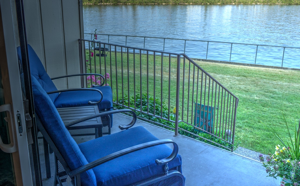 A private deck features two blue chairs facing Lake Pend Oreille, providing a serene spot to relax. The grassy area stretches toward the water, and a railing ensures safety. Colorful plants add a touch of nature to the setting.