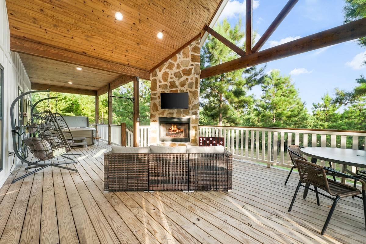 A covered wooden deck features a stone fireplace, providing a cozy focal point. A circular swing chair and a dining table with matching chairs are set against a backdrop of lush trees. Natural light enhances the inviting space, ideal for relaxation.