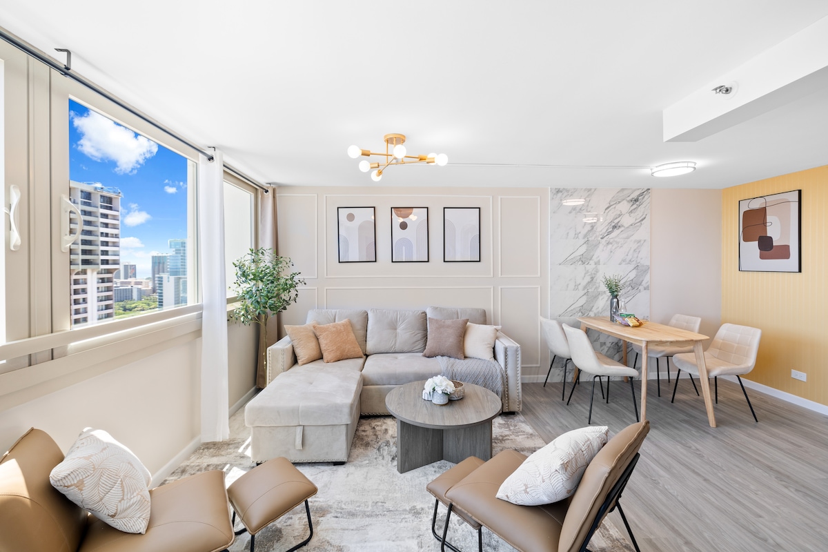 A bright living area with large windows offers views of the city skyline. A neutral-toned sectional sofa and two armchairs surround a round coffee table. A dining table with chairs is situated against a marble accent wall, complemented by framed artwork on the adjacent wall.