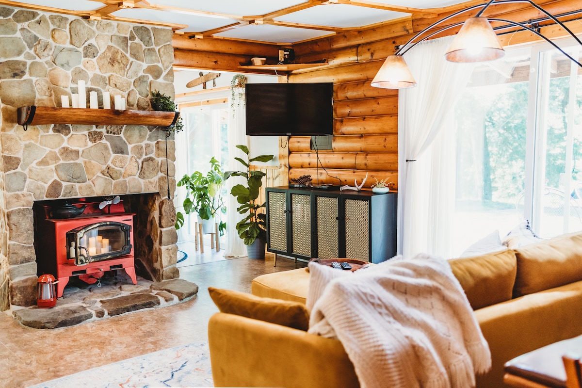 A cozy living area features a stone fireplace with a red wood stove, surrounded by log walls. A large window allows natural light to fill the space, while a plush sofa and decorative rug add comfort. Green plants enhance the inviting atmosphere.