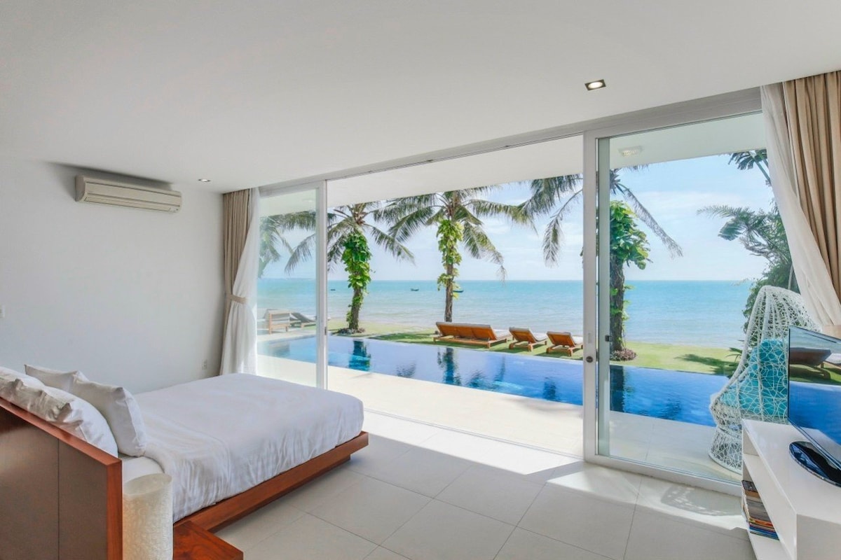 A modern bedroom features a spacious bed facing large glass doors that open to a stunning beach view. The serene outdoor area includes a pool and sun loungers surrounded by palm trees, allowing for direct access to the beach.