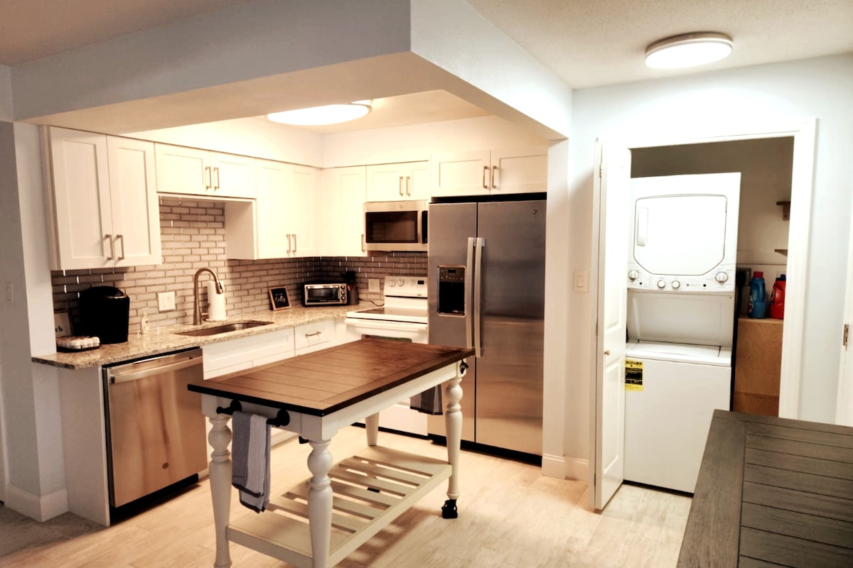 The modern kitchen features white cabinetry and stainless steel appliances, including a refrigerator and microwave. A center island with a wooden top provides additional workspace. A stacked washer and dryer are visible in a separate area, enhancing convenience.