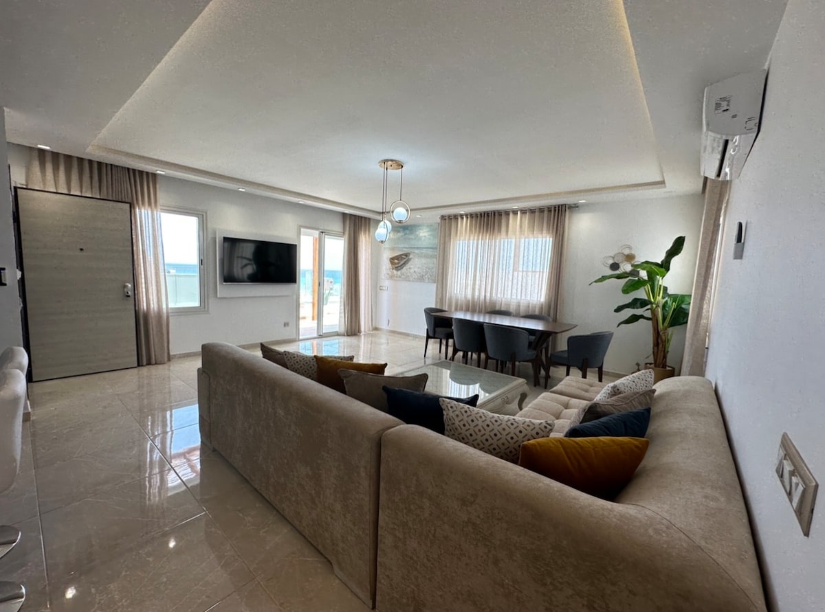 A bright and airy living space features a beige sofa adorned with colorful cushions, facing a wall-mounted television. A dining table is positioned nearby, surrounded by dark chairs. Large windows allow natural light to fill the room, complemented by elegant light fixtures.