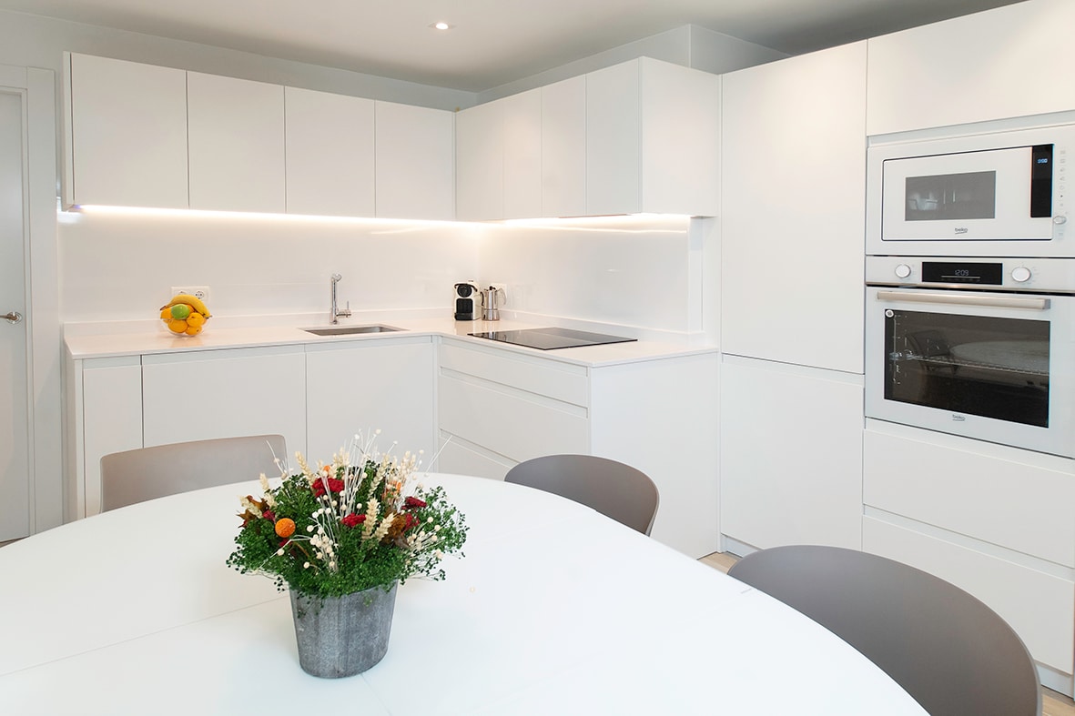 A modern kitchen features sleek white cabinetry and a minimalist design. A center dining table is surrounded by several chairs. Fresh flowers in a vase add a touch of color to the space. Integrated appliances are visible, offering a clean and functional cooking area.