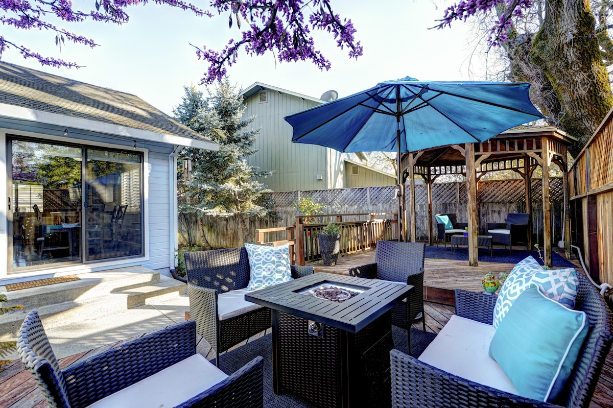 An outdoor seating area is furnished with plush chairs arranged around a square fire table. A large umbrella provides shade over the space. In the background, a gazebo offers additional seating options, surrounded by wooden fencing and vibrant greenery.