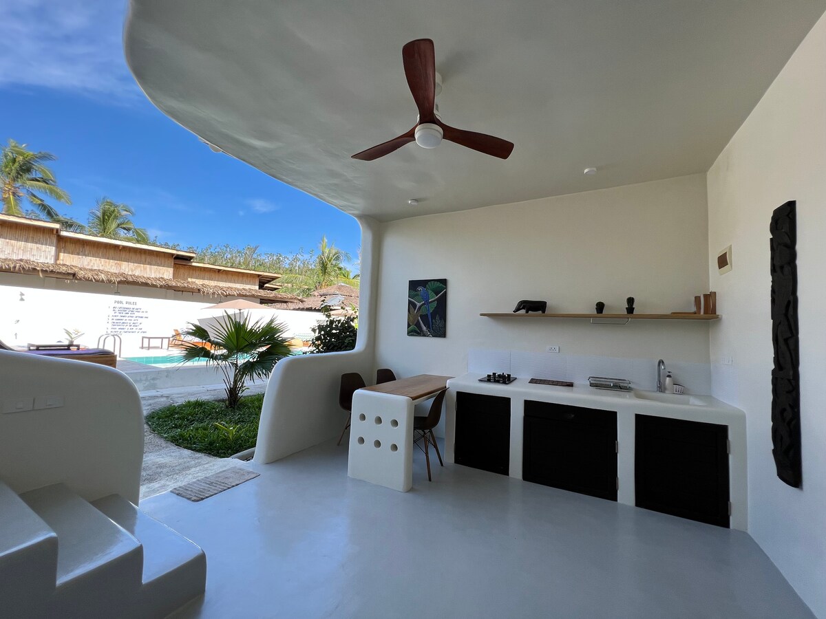 An open outdoor kitchen space is presented, featuring a modern design with a countertop, sink, and stovetop. A wooden dining table and chairs are arranged nearby. Lush greenery and a swimming pool can be seen in the background under clear blue skies.