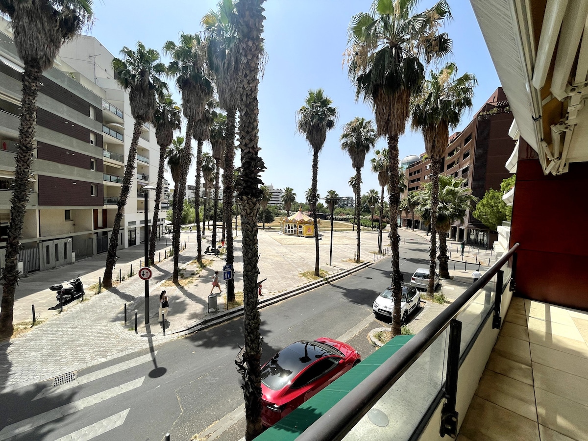 A balcony view showcases a palm-lined street, leading towards a plaza with colorful structures. The scene captures a sunny day, with vehicles parked along the side of the road. A prominent mix of greenery and architecture is visible from the first floor.