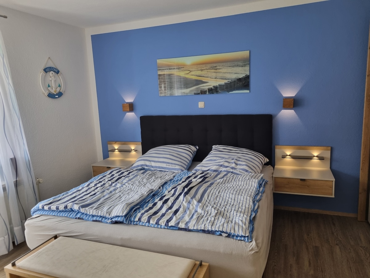 A cozy bedroom features a spacious bed with striped bedding against a blue accent wall. Two bedside lamps provide soft lighting, and there are minimalist wooden nightstands on either side. A decorative piece depicting a serene beach scene hangs above the bed.
