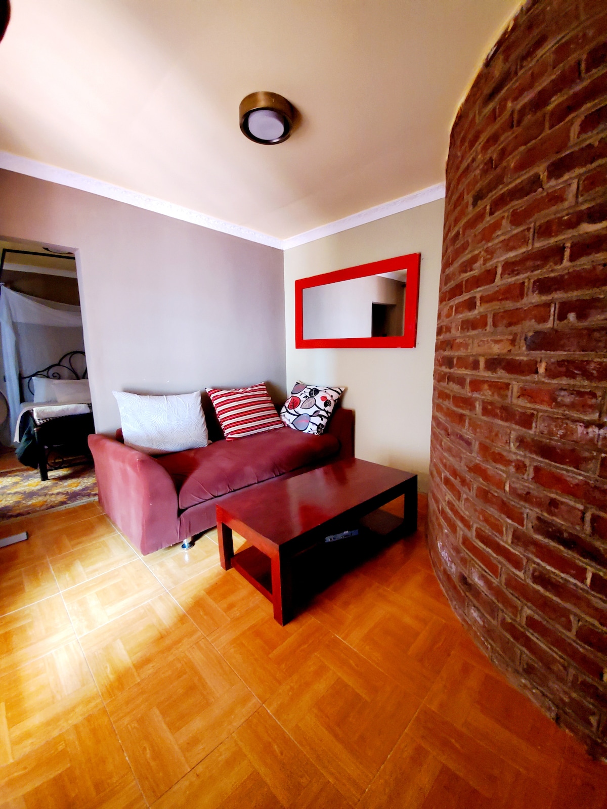 A cozy sitting area features a maroon sofa with decorative pillows and a small wooden coffee table. A brick accent wall adds texture, while soft light illuminates the space. A doorway leads to a bedroom in the background, enhancing the inviting atmosphere.