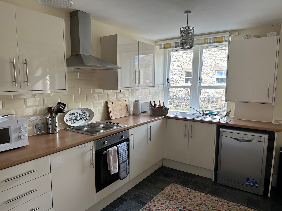 A compact kitchen features modern cabinetry and a wooden countertop. Essential appliances include a microwave, dishwasher, and oven setup. A decorative rug lies on the floor, and a window offers natural light, revealing views of the outdoor area. Utensils and cookware are arranged neatly.