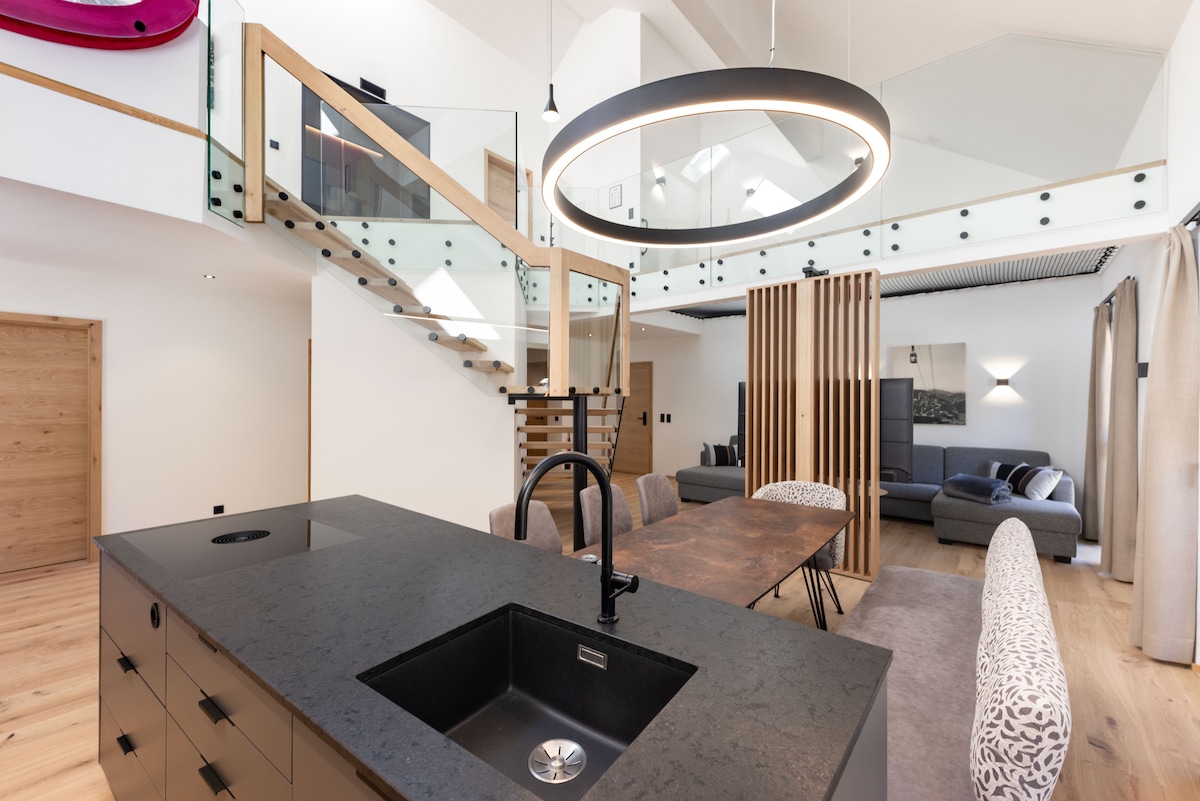 An open-plan area showcases a modern kitchen island with a black sink, complemented by a wooden dining table at the center. A staircase leads to a loft area, while a spacious living area is visible in the background, separated by wooden slats.