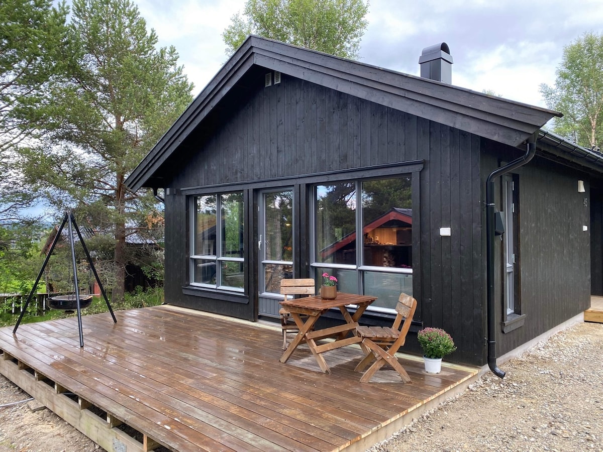 A modern black wooden cabin features large windows that provide views of the surrounding nature. The deck contains a wooden table with two chairs, ideal for outdoor dining. A swing set can be seen nearby, adding to the family-friendly atmosphere.