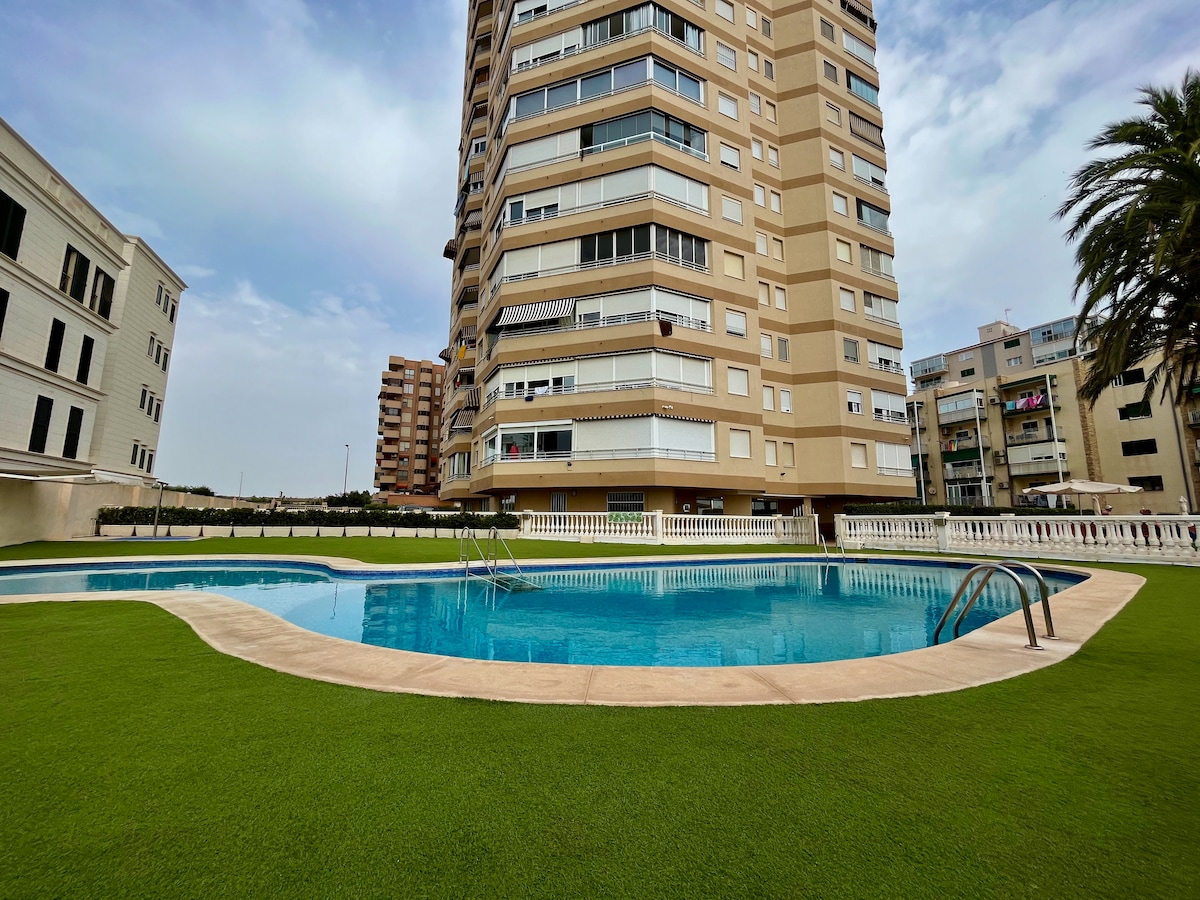 A multi-story building is shown, featuring a swimming pool surrounded by well-maintained green grass. The pool area is equipped with steps for easy access, and palm trees are visible nearby, contributing to the open outdoor space.