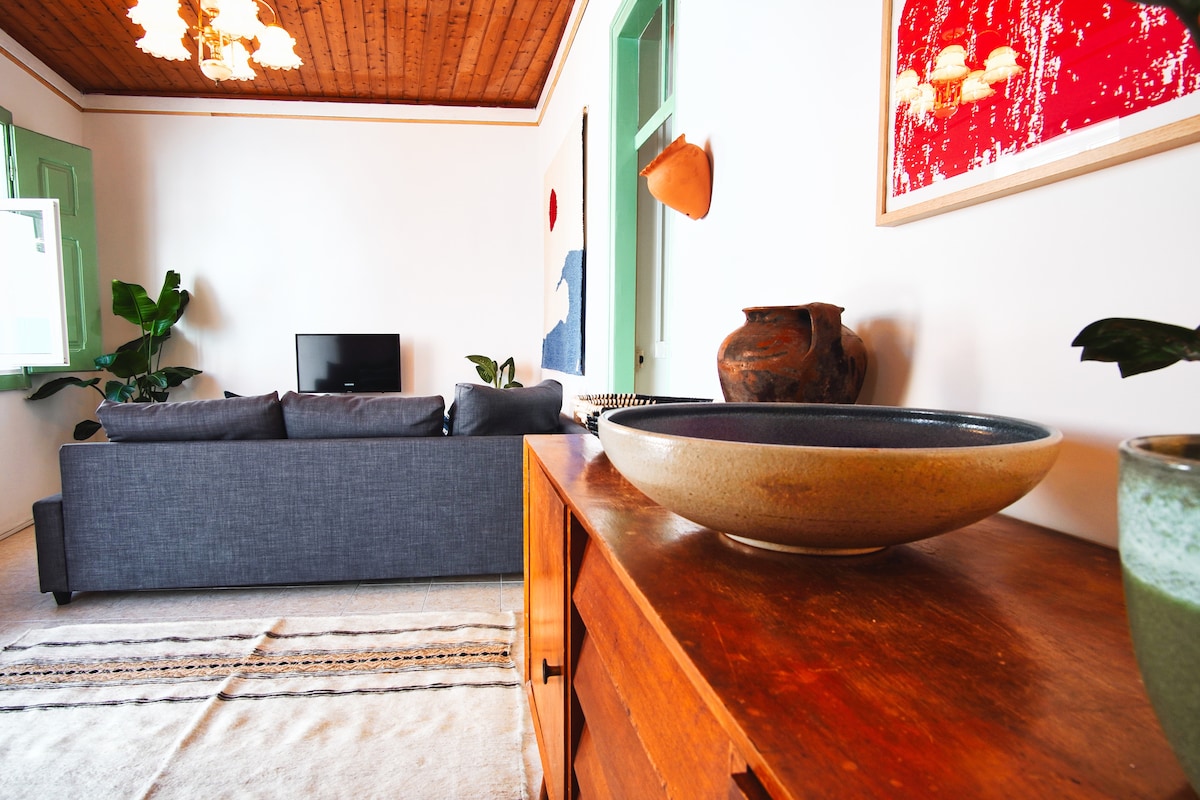 A cozy living area features a comfortable gray sofa positioned across from a television. Decorative plants add a touch of greenery, while a wooden sideboard displays a large bowl and a ceramic pot, creating a welcoming and relaxed environment.