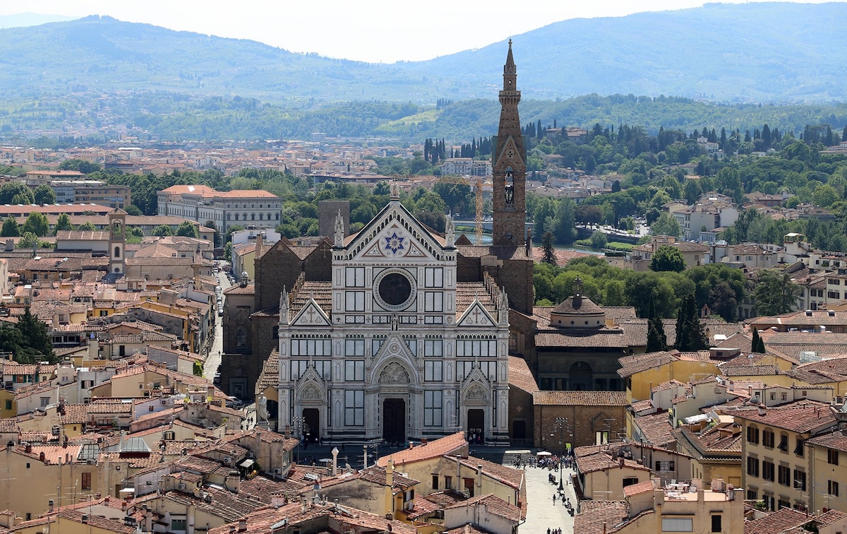 The image captures a panoramic view of Florence, featuring the Basilica di Santa Croce's intricate façade and tall bell tower. Surrounding rooftops and the distant hills enhance the historical charm of the city, with greenery visible in the background.