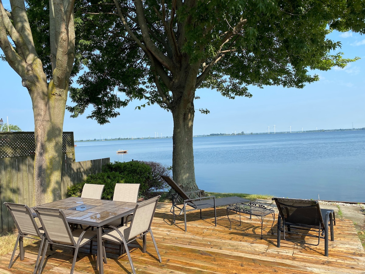 An outdoor seating area features a large table surrounded by multiple chairs, positioned under the shade of two large trees. A pair of lounge chairs are placed nearby, offering views of the calm water of the bay and beyond.
