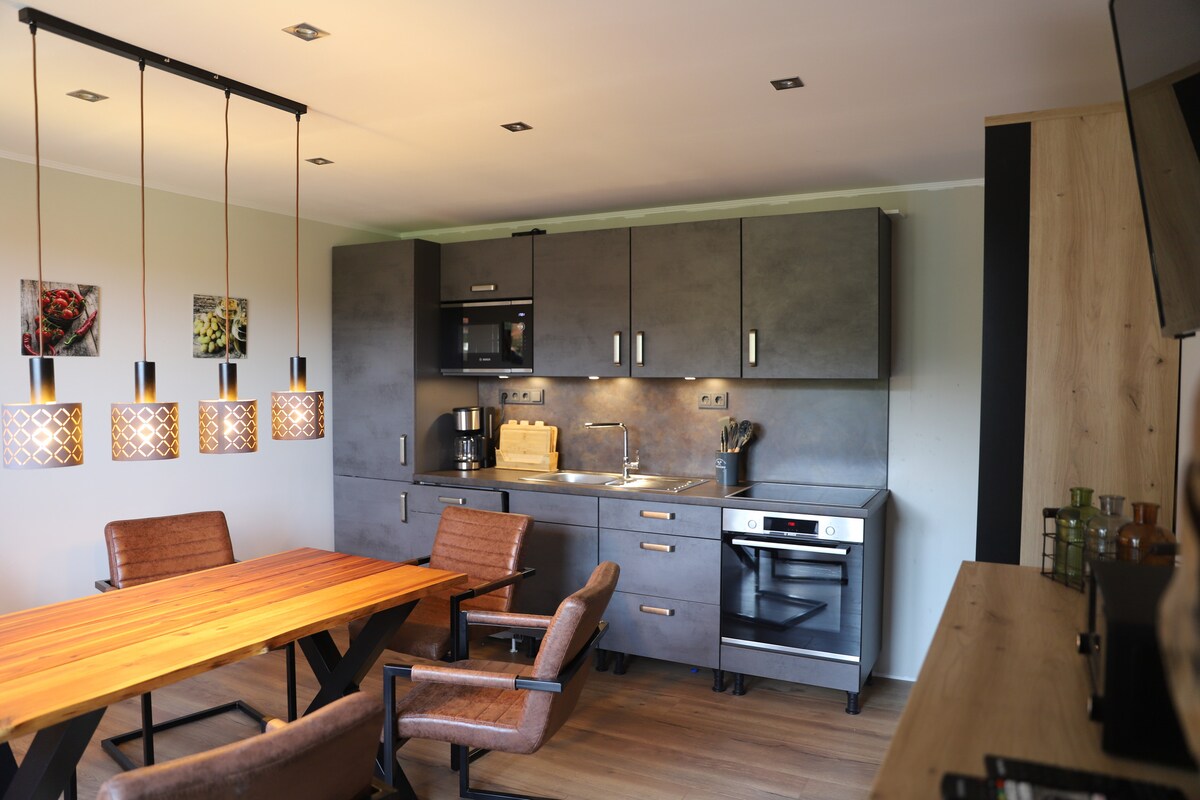 A modern kitchen area is visible, featuring sleek dark cabinetry and stainless steel appliances. A wooden dining table, accompanied by four brown chairs, provides ample seating. Unique pendant lighting hangs above the table, creating a sophisticated ambiance.