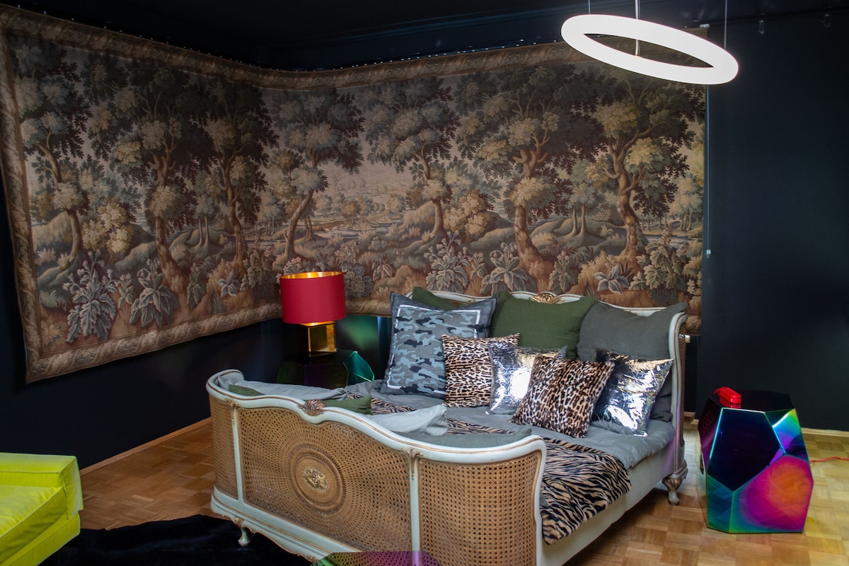 A seating area is featured with a vintage sofa adorned with various cushions in animal prints and textures. A pendant light hangs above, and a large, ornate wall tapestry depicting a natural scene provides a backdrop. A colorful side table adds a contemporary touch.