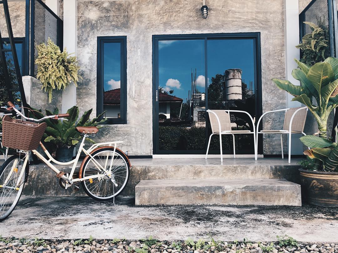 The entrance to the property features a wide concrete step leading to a patio area. Two white chairs are positioned in front of large glass doors, reflecting the scenery outside. A bicycle with a basket is parked nearby, surrounded by greenery.