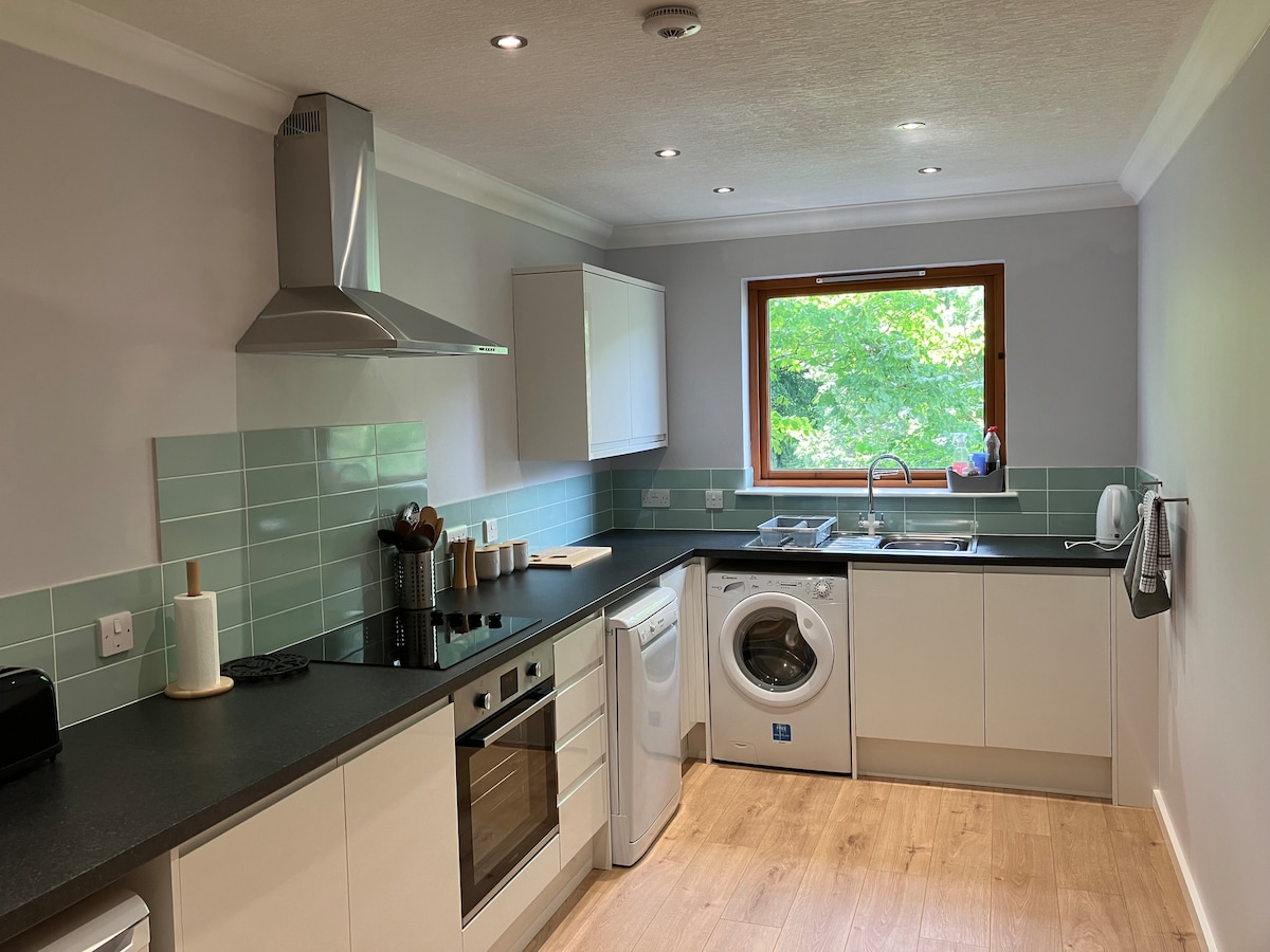 The kitchen area features modern appliances, including an oven, stovetop, microwave, and washing machine. Light wood flooring complements the sleek cabinetry, while a window provides natural light and views of greenery outside. Counter space is available for meal preparation.