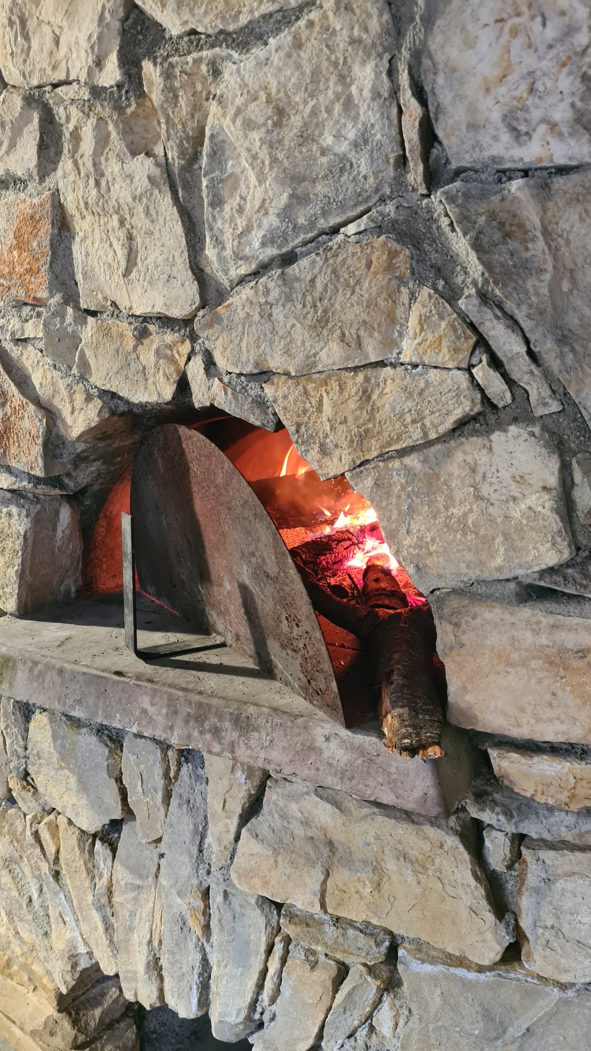 A rustic stone fireplace displays a warm, glowing fire within its spacious archway. The detailed stonework emphasizes the craftsmanship, while a log rests beside the flames, enhancing the inviting atmosphere of the area.