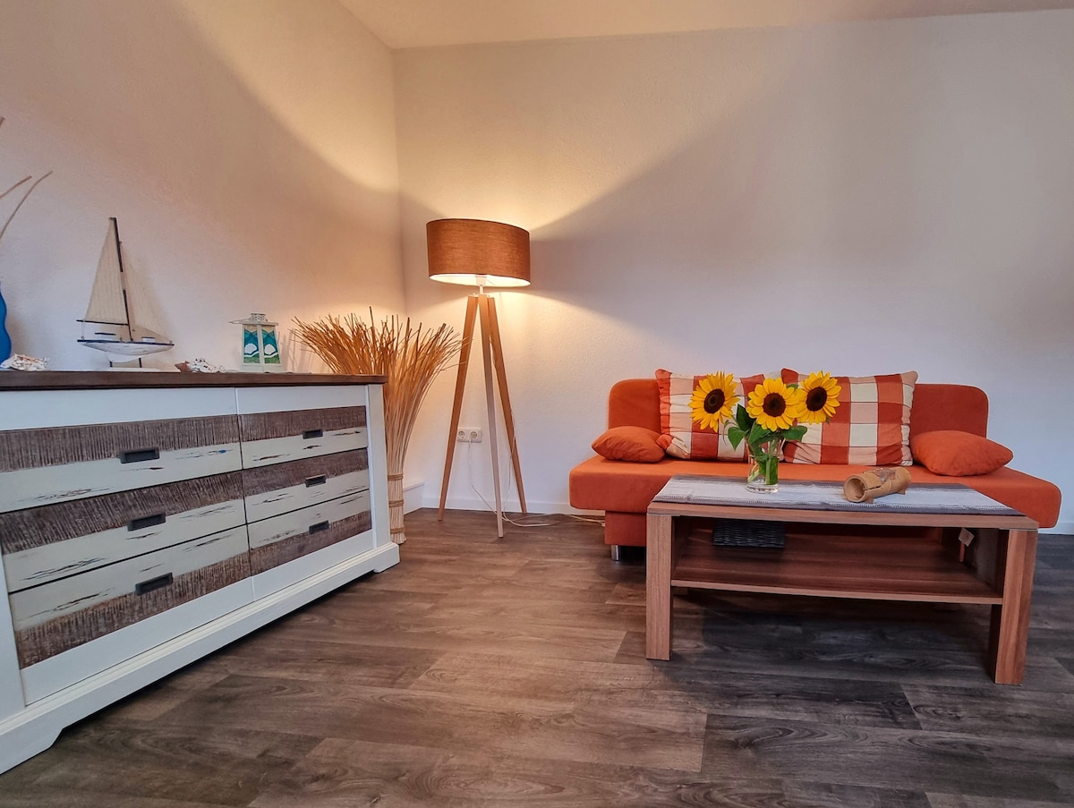 A living area features a vibrant orange sofa adorned with decorative pillows, positioned next to a light wooden coffee table. A vase of sunflowers adds a cheerful touch, while a tall floor lamp provides warm lighting. A decorative cabinet is situated against the wall, enhancing the space.