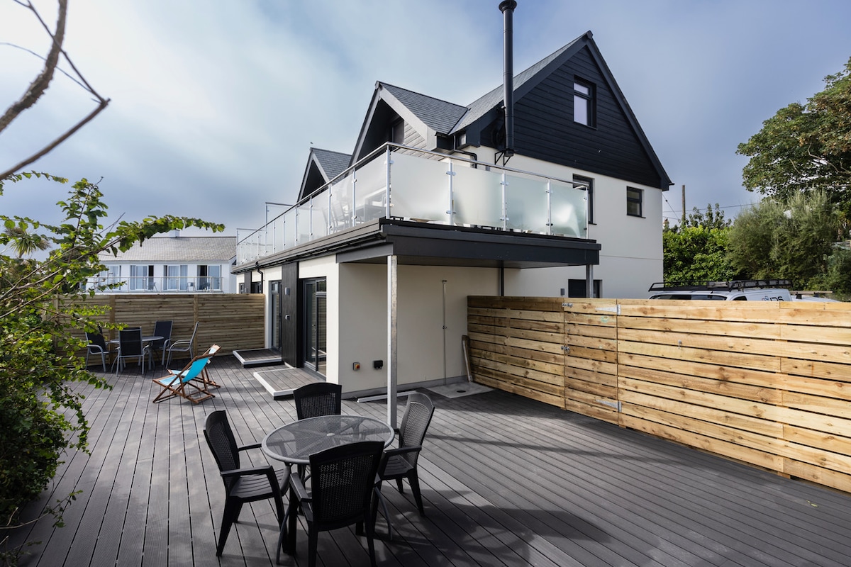 The exterior features a modern semi-detached house with a large deck. A glass balcony overlooks the outdoor seating area, which includes a round table and several black chairs. A wooden fence provides privacy, while a few lounge chairs are positioned along the deck.