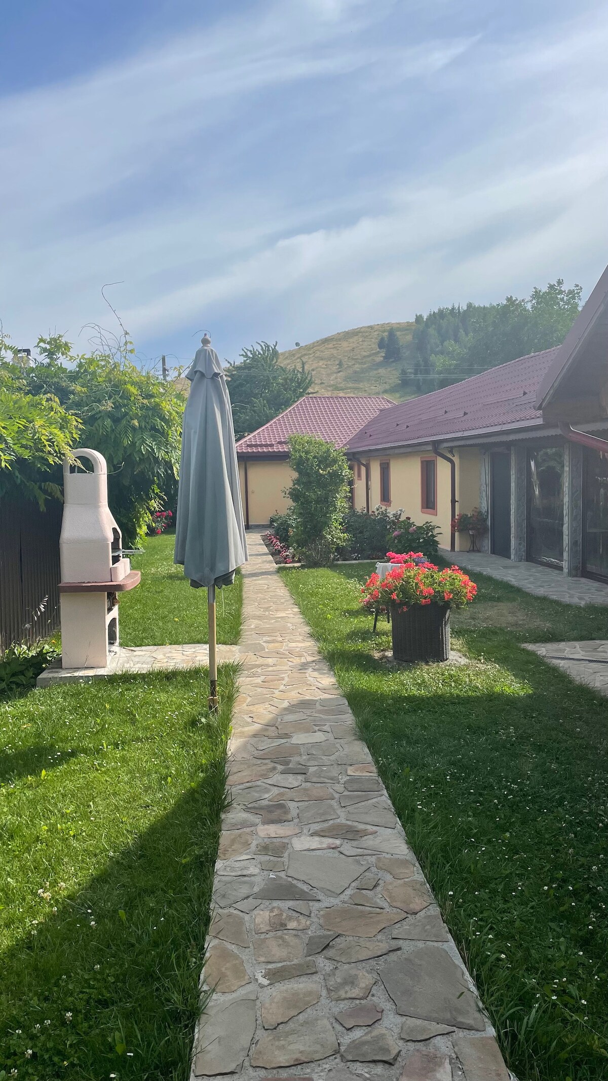 A stone pathway winds through a green garden, bordered by flowering plants and a tall umbrella. The yard features a barbecue grill on one side and buildings with red roofs in the background, surrounded by gentle hills and trees.