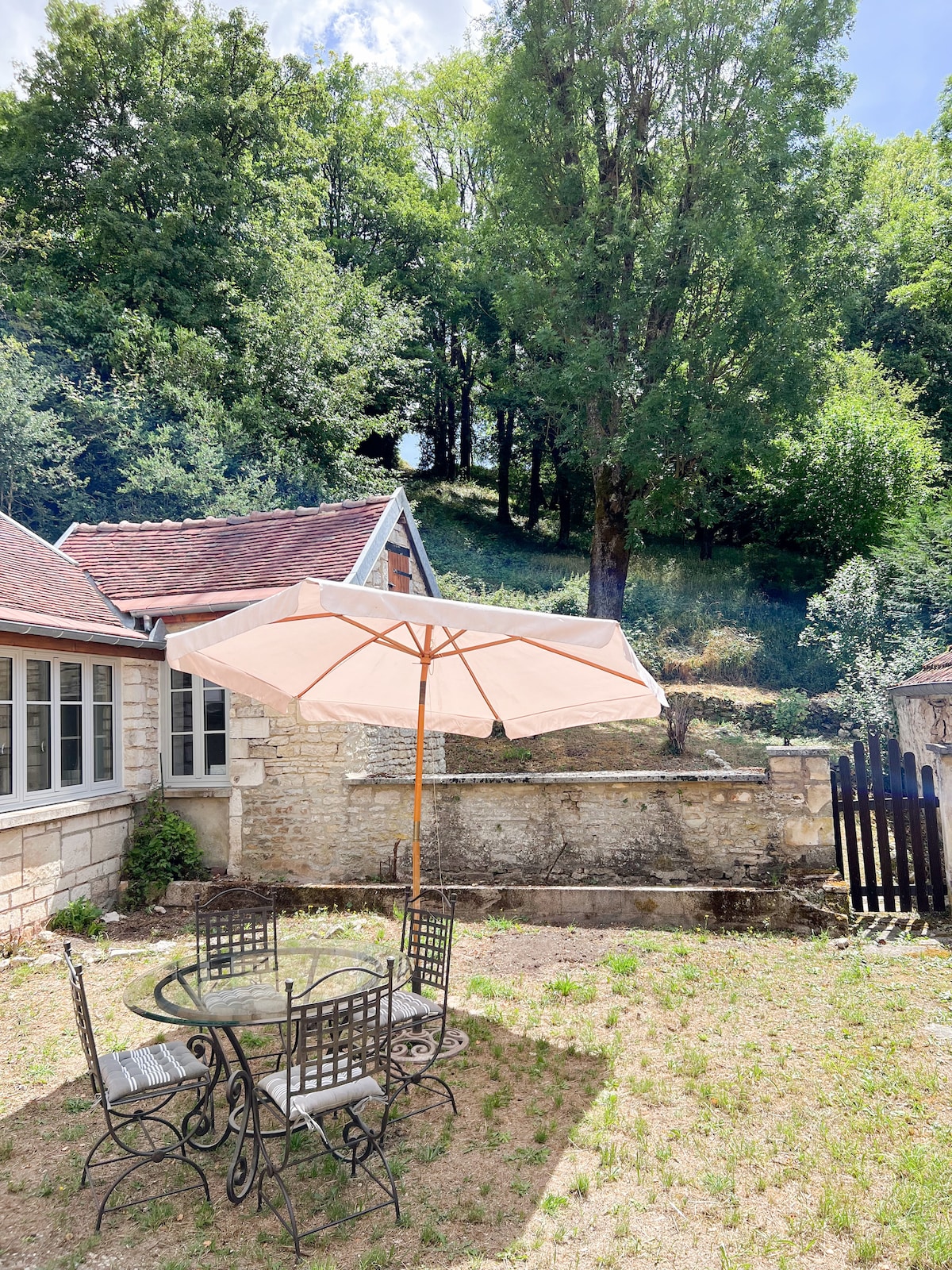 An outdoor seating area features a round glass table surrounded by four metal chairs. A large umbrella provides shade, and the backdrop includes a stone wall and a lush, wooded area with tall trees. The space is peaceful and inviting.