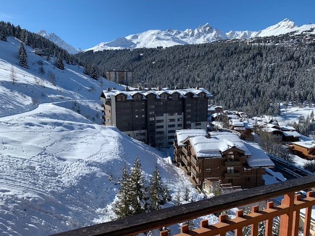 A panoramic view showcases snow-covered mountains in the background, with a multi-story building set against the hillside, surrounded by evergreen trees and a snow-blanketed landscape. The scene provides a sense of tranquility and winter beauty, emphasizing the natural surroundings of the area.