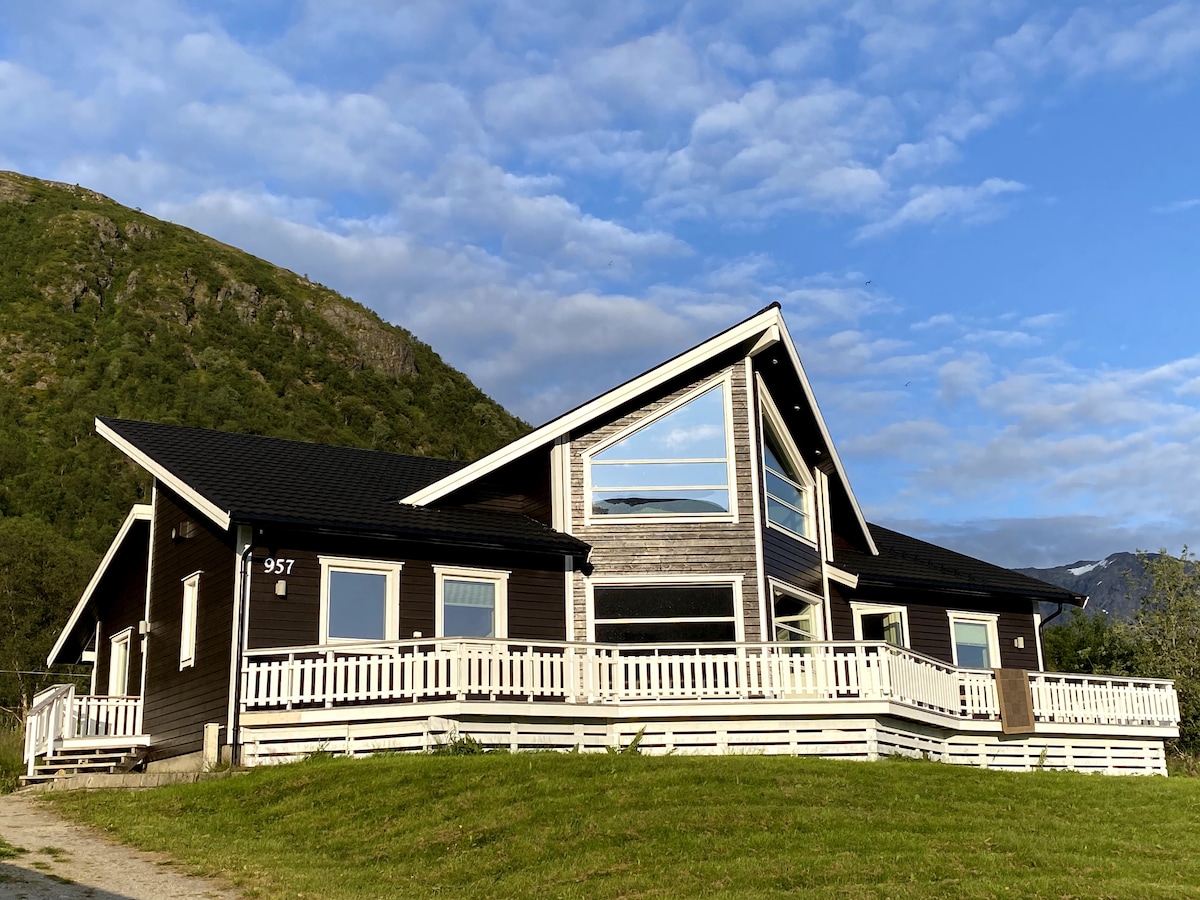 The exterior of a spacious holiday home features a steeply pitched roof and multiple large windows, allowing natural light to fill the interiors. A broad deck is surrounded by a white railing, providing scenic views of the surrounding mountains and expansive sky.
