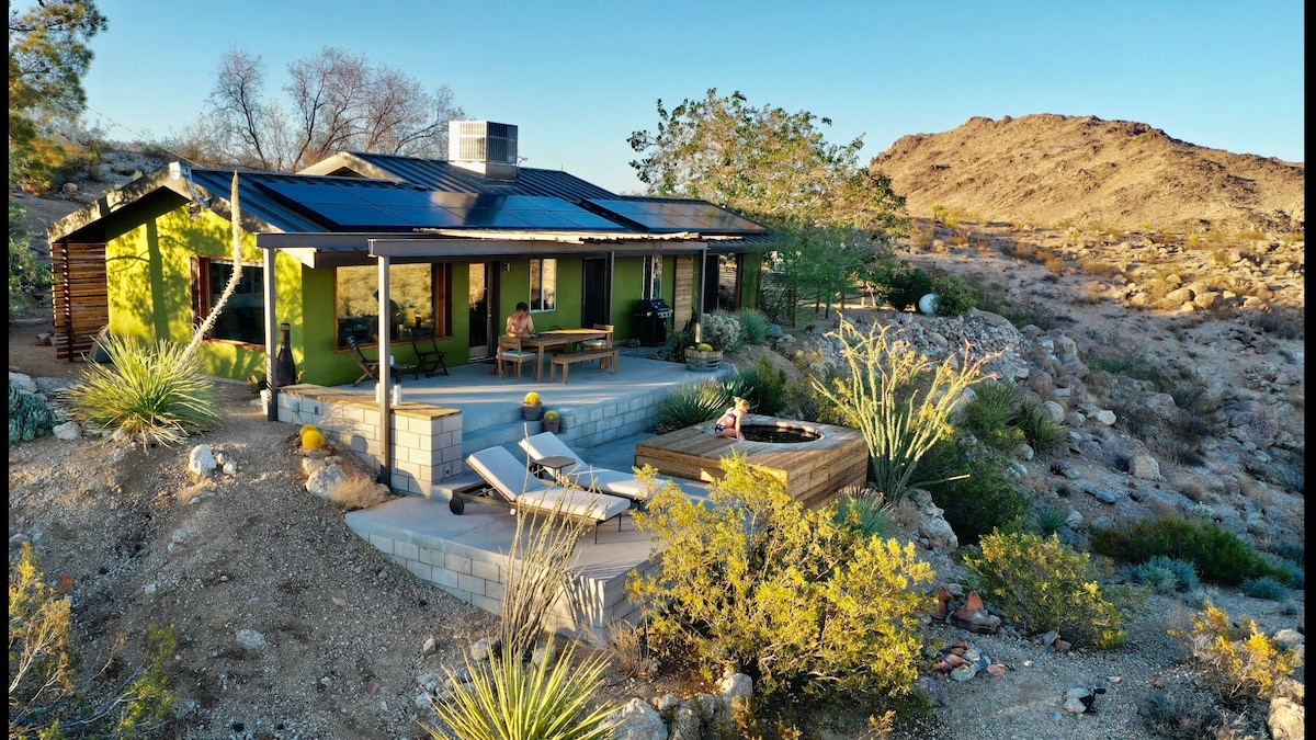 The retreat features an eco-friendly design with solar panels on the roof, surrounded by native desert vegetation. An outdoor seating area and hot tub are present on a spacious terrace, offering views of the rocky landscape and nearby mountains.