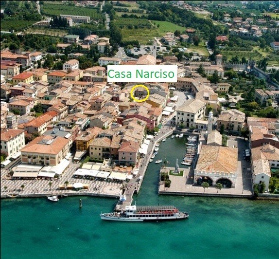 An aerial view of a picturesque town by the Garda Lake is presented, featuring a mix of colorful rooftops and structures lining the waterfront. A boat is docked in the harbor, and green hills can be seen in the background, enhancing the scenic landscape.